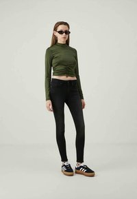 Mulher usando óculos de sol escuros, blusa cropped de manga longa em verde-oliva com franzidos, jeans skinny pretos e ténis pretos com riscas brancas e solas de borracha.