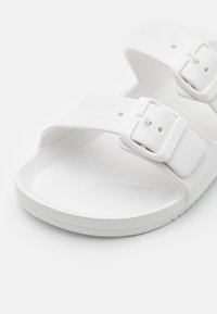 Bela slip-on sandala iz gladkega sintetičnega materiala, ki ima dva nastavljiva traka z belimi zaponkami in teksturiran podplat.