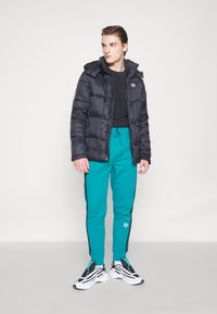 Sort pufferjakke med hætte, lynlås foran og sidelommer; teal træningsbukser med sorte side-striber; hvide og sorte sneakers.