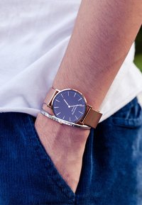 Gold mesh wristwatch with a blue face, featuring thin white hour markers. Silver bracelet visible alongside it, worn on a bare arm.