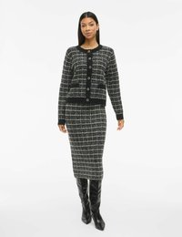 Cardigan en tweed noir et blanc avec col rond, fermeture à boutons et poches, associé à une jupe assortie et des bottes noires ajustées à hauteur de genou.