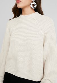 Frau trägt einen cremefarbenen Strickpullover mit strukturiertem Muster und großen schwarz-weißen kreisförmigen Ohrringen.