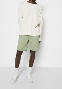 Persona che indossa una maglietta a maniche lunghe color crema a coste, pantaloni corti verde chiaro fino al ginocchio, calze bianche e scarpe da tennis bianche, in piedi contro un muro bianco.