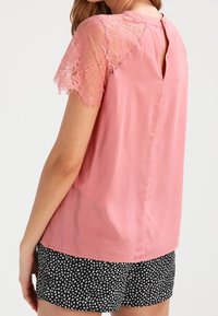 Modell som bär en rosa blus med spetsärmar och en nyckelhålsöppning bak, i kombination med svarta shorts med ett vitt blommönster.