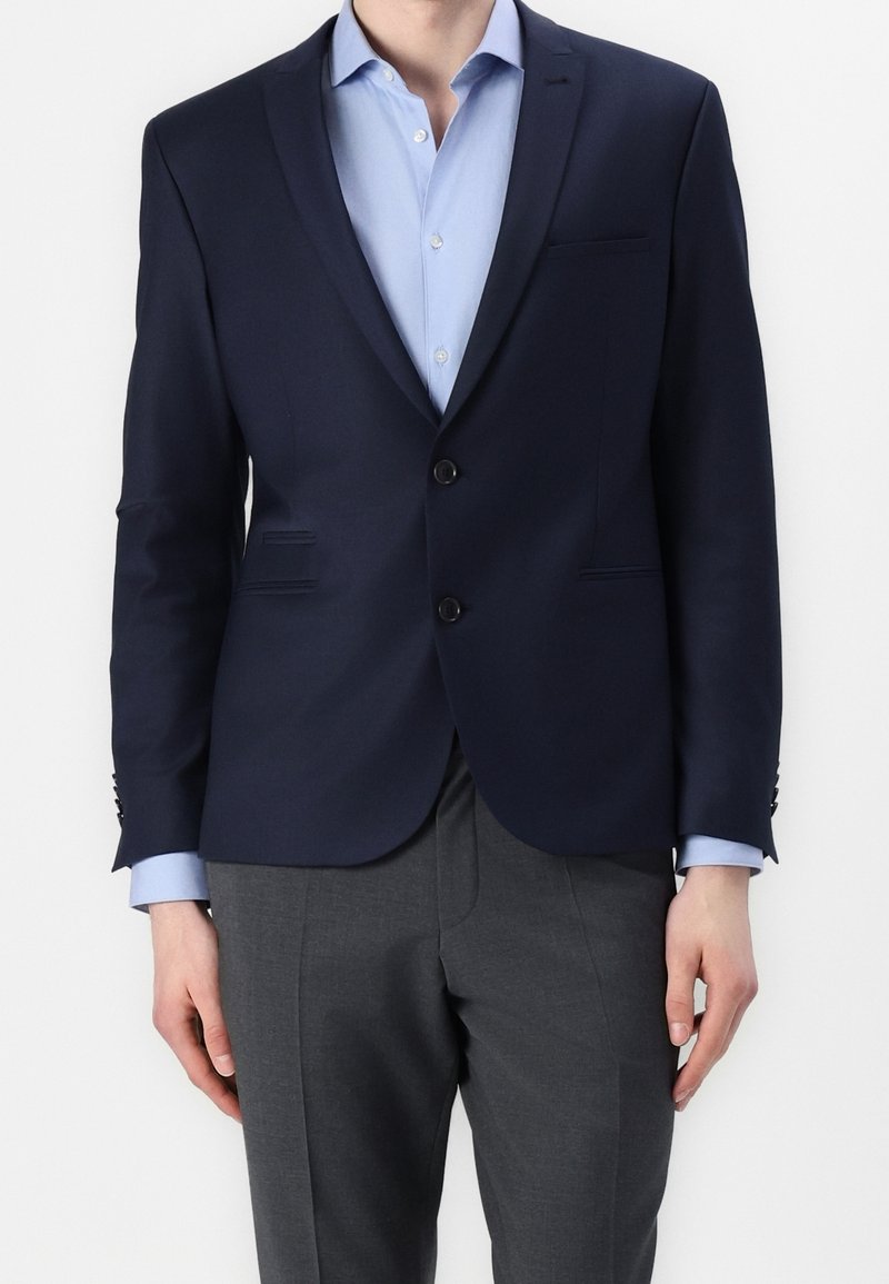 Granatowa marynarka z klapami notch, zapięciem na dwa guziki i bocznymi kieszeniami. W połączeniu z jasnoniebieską koszulą i szarymi spodniami. Gładka tekstura.