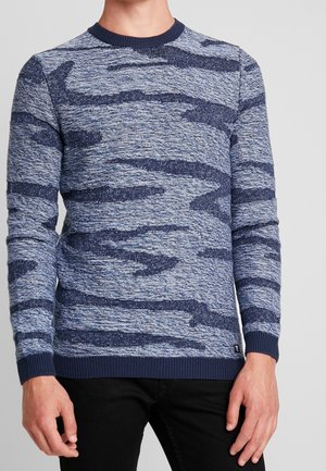 Un homme portant un sweat-shirt à manches longues col rond avec un motif texturé abstrait bleu et marine, et un pantalon noir.
