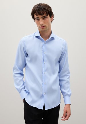 Camisa de manga larga azul claro con un pequeño patrón de cuadros, cuello abotonado y botones blancos. La tela tiene una textura suave. Combinada con pantalones negros.