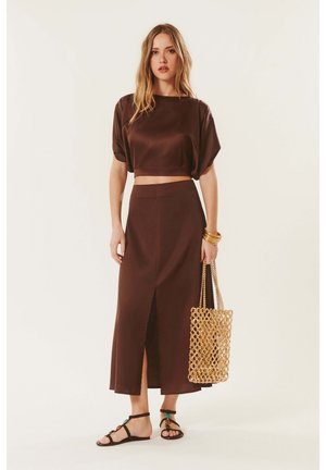 Femme debout portant un haut court marron et une longue jupe assortie avec une fente à l'avant, tenant un sac tressé beige et portant des sandales noires.