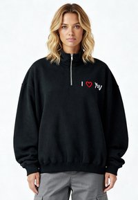 Sweat-shirt en polaire noire avec un col montant. Il comporte une fermeture à glissière sur la moitié et un message brodé « I ❤️ YOU » en rouge et blanc sur le devant.