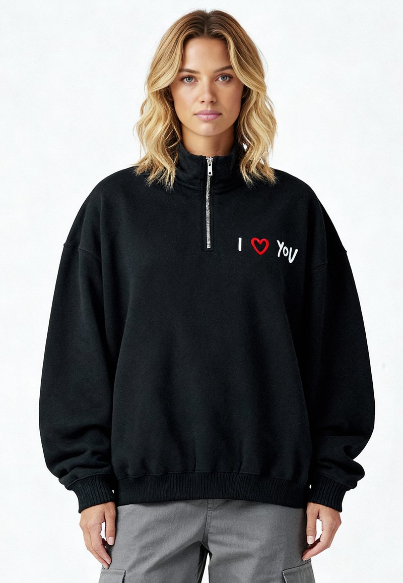 Sweat-shirt en polaire noire avec un col montant. Il comporte une fermeture à glissière sur la moitié et un message brodé « I ❤️ YOU » en rouge et blanc sur le devant.