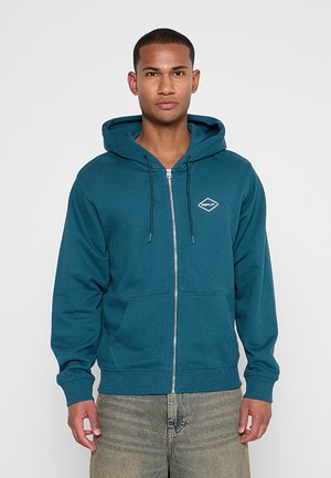 Mann trägt einen türkisfarbenen Kapuzenpullover mit Reißverschluss, Fronttaschen und „Replay“-Logo auf der Brust, kombiniert mit hellgrauen Jeans, steht vor weißem Hintergrund.