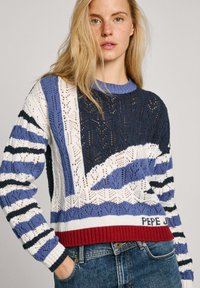 Pull en tricot de couleur marine, bleue et blanche, avec un design à rayures et une texture en maille ouverte. Ourlet rouge avec le nom de la marque en bas.