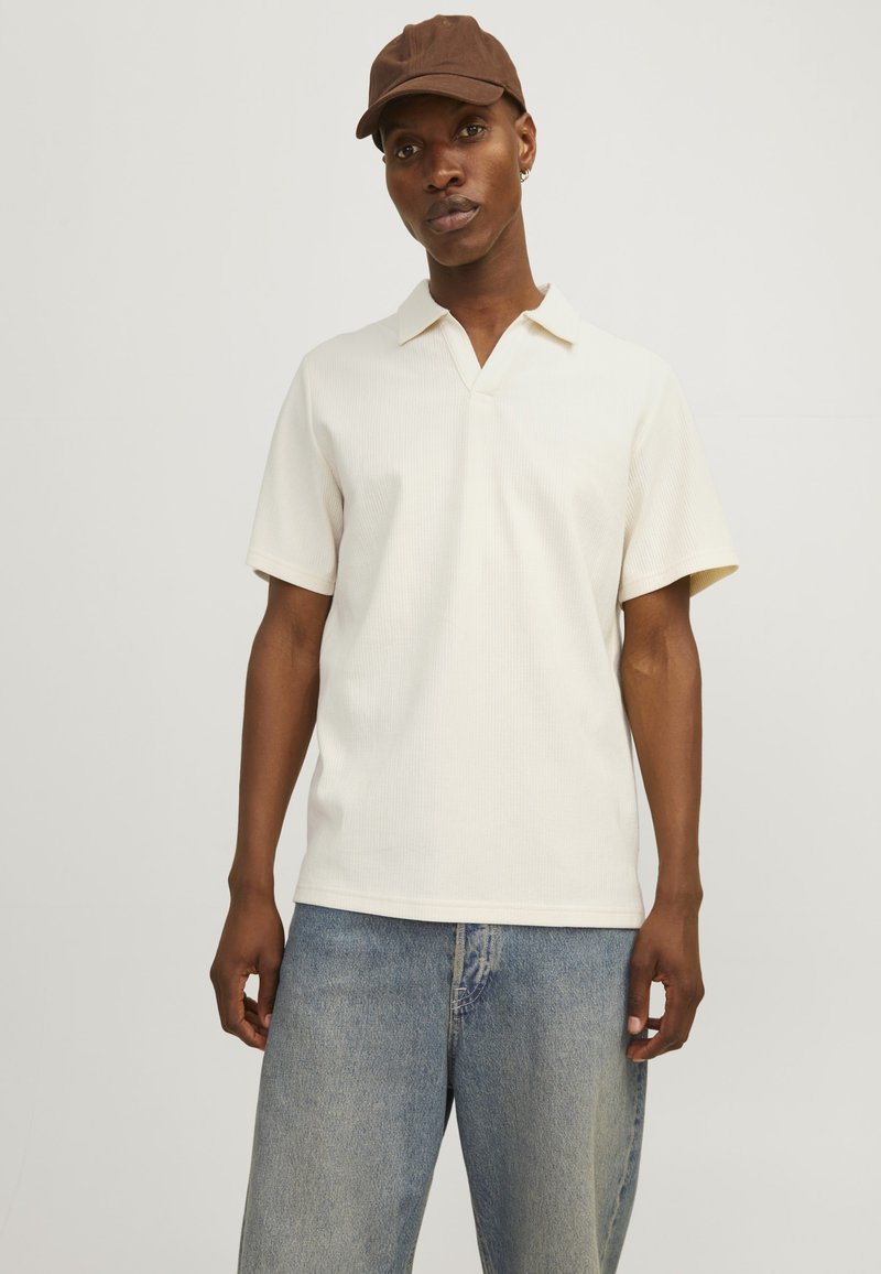 Camisa polo de manga corta en color crema con textura acanalada y cuello en V, combinada con pantalones cortos de mezclilla de tiro alto en azul claro. Lleva puesta una gorra marrón.