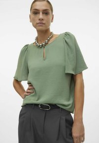 Top verde chiaro a maniche corte con finitura testurizzata e scollatura a occhio di pernice, abbinato a pantaloni grigio scuro e una collana di perline argentate.