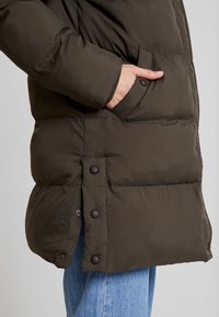 Brun polstret frakke med en quiltet tekstur, sidelommer og brune trykknapper ved kanten og manchetterne. Båret over blå denim.