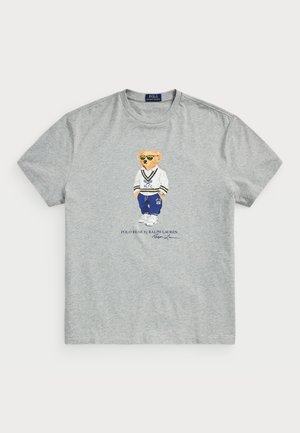 Szara koszulka z okrągłym dekoltem z misiem w okularach przeciwsłonecznych, białym swetrze, niebieskich spodniach i białych trampkach, z napisem „Polo Bear by Ralph Lauren” poniżej.