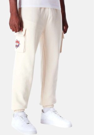 Joggers color crema con bolsillos laterales de cargo y diseños bordados. Ajuste slim, puños acanalados y tela de mezcla de algodón. Combinados con zapatillas blancas.
