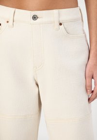 Beige cotton denim pants with a high waist, five-pocket design, and brass button closure. Features stitching accents and a straight-leg cut.