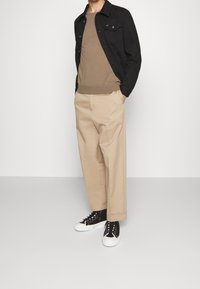 Schwarze Jacke mit Knöpfen, beiger Strickpullover, bequeme beige weit geschnittene Hose und schwarze Sneakers mit weißen Sohlen. Einfaches, lässiges Outfit.