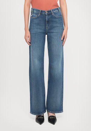 Rechthoekige blauw-wijde jeans gedragen met zwarte puntige loafers door een staand persoon tegen een witte achtergrond.