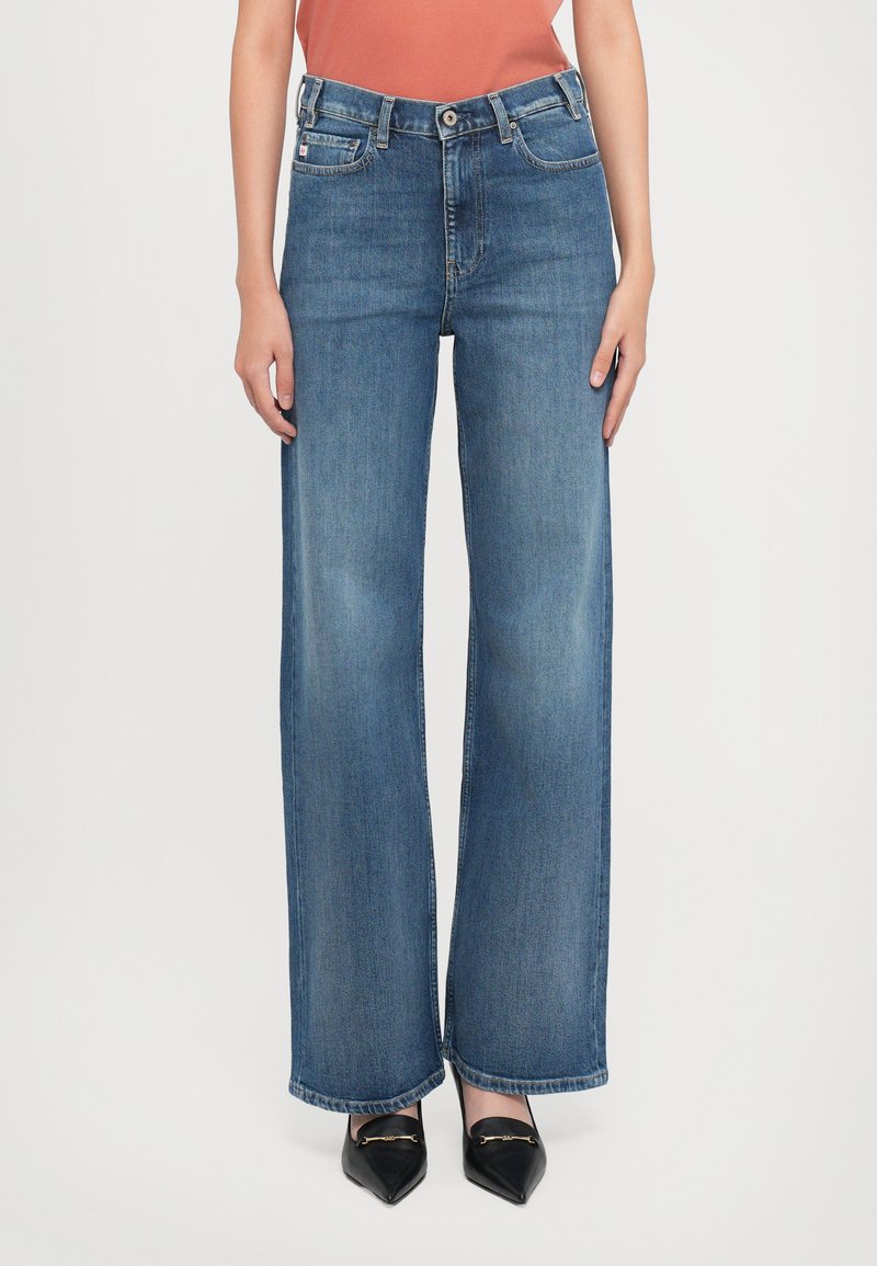Jeans azuis de perna larga e reta, usados com mocassins pretos de bico, em uma pessoa em pé diante de um fundo branco.