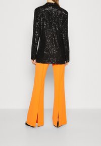 Blazer negra con lentejuelas y mangas largas, combinada con pantalones acampanados naranja brillante con aberturas laterales, sobre un fondo liso gris claro.