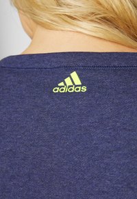 Personne blonde portant un t-shirt bleu foncé avec un petit logo Adidas jaune fluorescent sur la partie haute du dos, au niveau de la nuque.