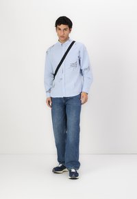 Light blue button-up shirt with black line drawings, paired with blue denim jeans. The model carries a black crossbody bag and wears sneakers.