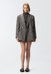 Sivi tweed oversize sako s zlatnim gumbima, s režanj kragnom, dva prednja džepa i teksturiranom tkaninom. Nošen preko tamne majice.