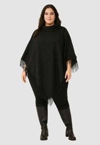 Poncho nero con scollatura a cappuccio, caratterizzato da dettagli frangiati lungo l'orlo, abbinato a leggings neri e stivali neri al ginocchio.