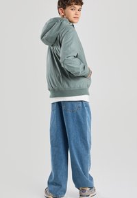 Vaaleanvihreä nailoninen bomber-takki, jossa on huppu, elastiset hihansuut ja helma. Päällä valkoinen paita ja yhdistettyinä löysät siniset farkut.