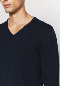 En närbild av en mans överkropp iklädd en mörkblå V-ringad tröja med en subtil logotyp på bröstet mot en ljus bakgrund.