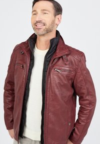 Burgunderfarbene Lederjacke mit schwarzem Innenfutter, ausgestattet mit Reißverschlusstaschen, hohem Kragen und detaillierter Näherei. Glatte Textur, moderner Schnitt.