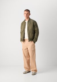Green bomber jacket with zippered pockets, paired with light pink cargo pants. Model wears beige and gray sneakers on a white background.