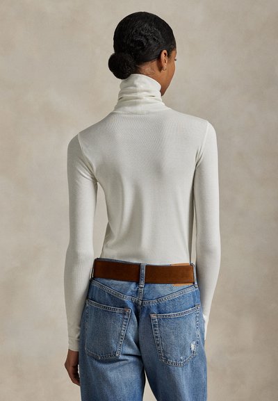 Sötét hajjal kontyba kötve, fehér garbót és kék farmert barna övvel viselő személy, háttal, egyszínű háttér előtt.