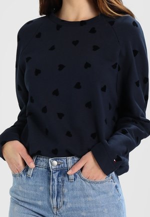 Femme portant une chemise bleu marine à manches longues avec des motifs de cœurs noirs rentrée dans un jean en denim bleu clair, les mains dans les poches.