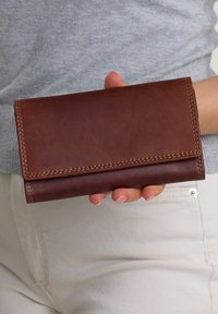 Braune Lederbörse mit einem Umschlagdesign, das Nähdetails und eine glatte Textur zeigt. In einer Hand vor einem hellen Hintergrund gehalten.