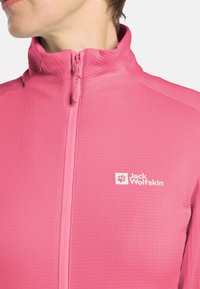 Veste zippée rose clair en tissu texturé, avec un col haut et le logo "Jack Wolfskin" en blanc sur la poitrine.