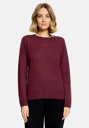 Femme aux cheveux blonds mi-longs portant un pull bordeaux à manches longues et un pantalon noir, debout devant un fond clair uni.