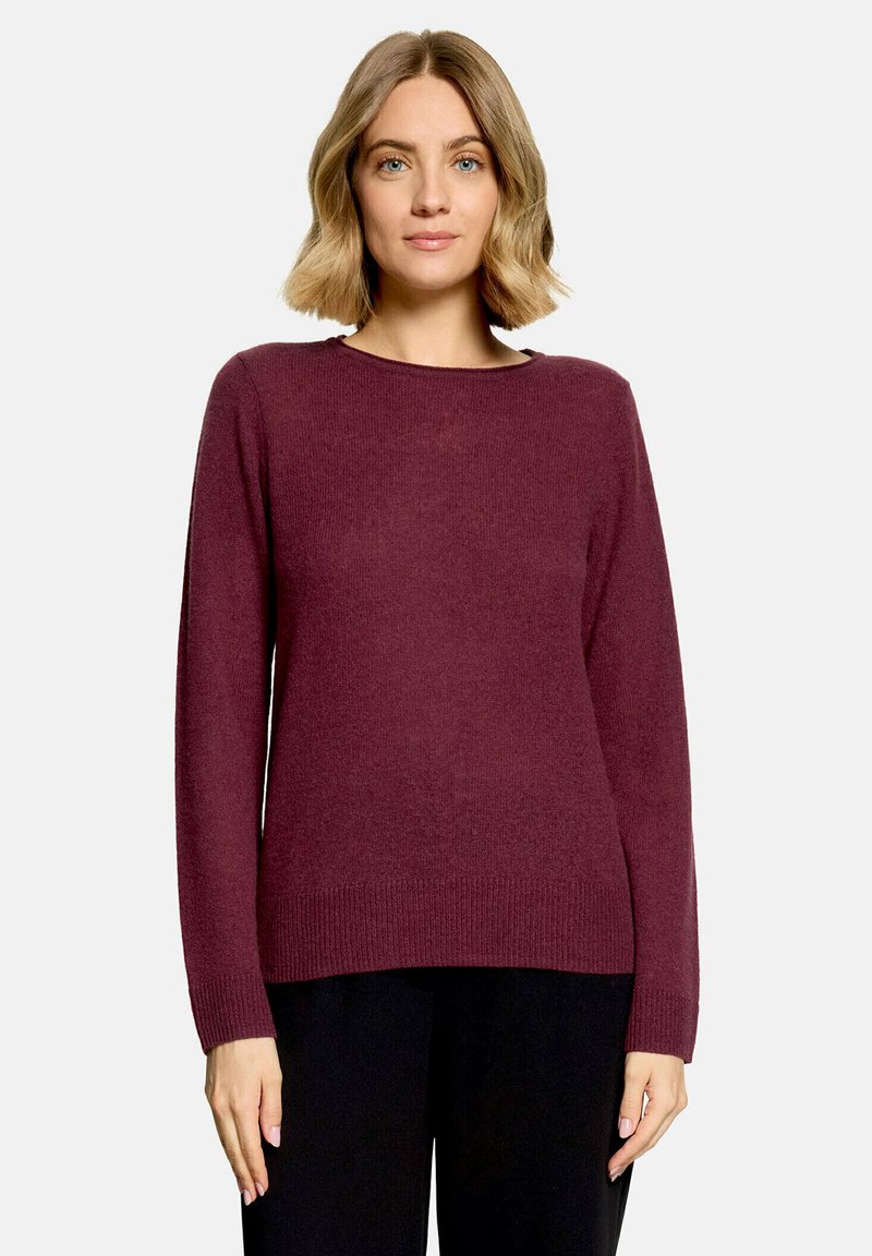 Femme aux cheveux blonds mi-longs portant un pull bordeaux à manches longues et un pantalon noir, debout devant un fond clair uni.