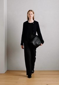 Schwarze Strickjacke mit Knöpfen, schwarzes Oberteil und weite Stoffhose. Das Model trägt eine große schwarze Lederhandtasche und geht über einen Holzboden.
