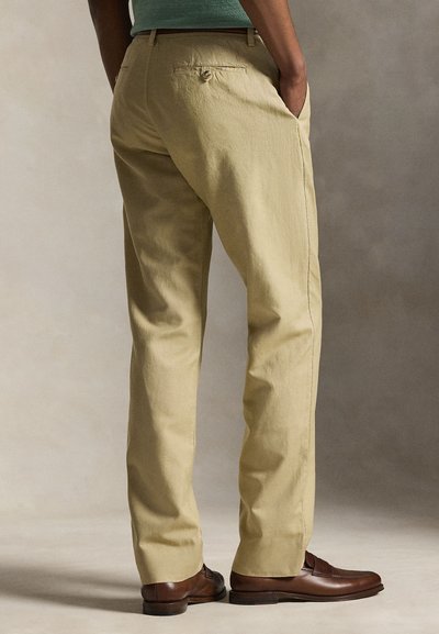 Mężczyzna w beżowych spodniach chino i brązowych skórzanych mokasynach, stoi z rękami w kieszeniach na neutralnym tle.