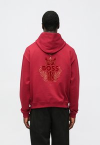 Felpa con cappuccio rossa con un elaborato disegno dorato e nero e la scritta "BOSS" sul retro. Realizzata in tessuto morbido e strutturato.
