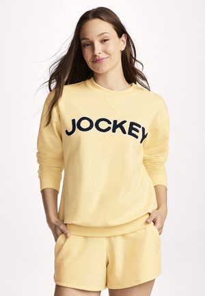Frau mit langen braunen Haaren, die einen gelben Pullover mit dem Schriftzug "JOCKEY" und passende gelbe Shorts trägt, die Hände in den Taschen, leicht lächelnd.