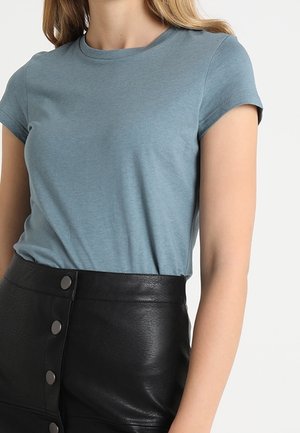 Kobieta ubrana w niebieski t-shirt z krótkim rękawem wpuszczony w czarną skórzaną spódnicę z guzikami z wysokim stanem, pokazana od szyi do bioder.