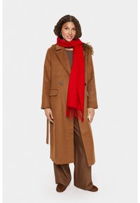 Kvinde med krøllet brunt hår iført en lang brun frakke, matchende bredbenede bukser, brune sko og et klart rødt tørklæde.