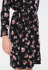 Černé květinové šaty s dlouhými rukávy, páskem v oblasti pasu a volným siluetou. Látka vykazuje červené a bílé květinové vzory.