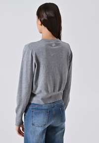 Donna con lunghi capelli castani, che indossa orecchini d'oro, un maglione grigio attillato con il logo sulla parte alta della schiena e jeans di denim blu, di spalle.