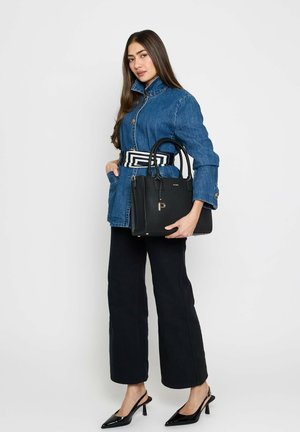 Young woman in denim jacket, black wide-leg pants, black heels, holding black handbag with geometric belt on white background.