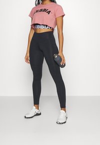 Leggings negros de tejido elástico, combinados con una camiseta corta rosa que muestra "NEBBIA" en letras negras, sosteniendo una botella de agua gris.